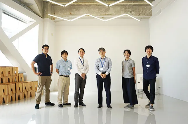 白い室内で7人のメンバーが横一列に並んで立っている集合写真